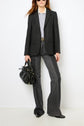 Person wearing an esprit tailleur with a black blazer, light gray button-up shirt, dark gray flared jeans, and black shoes, holding a black handbag, standing against a plain white background.