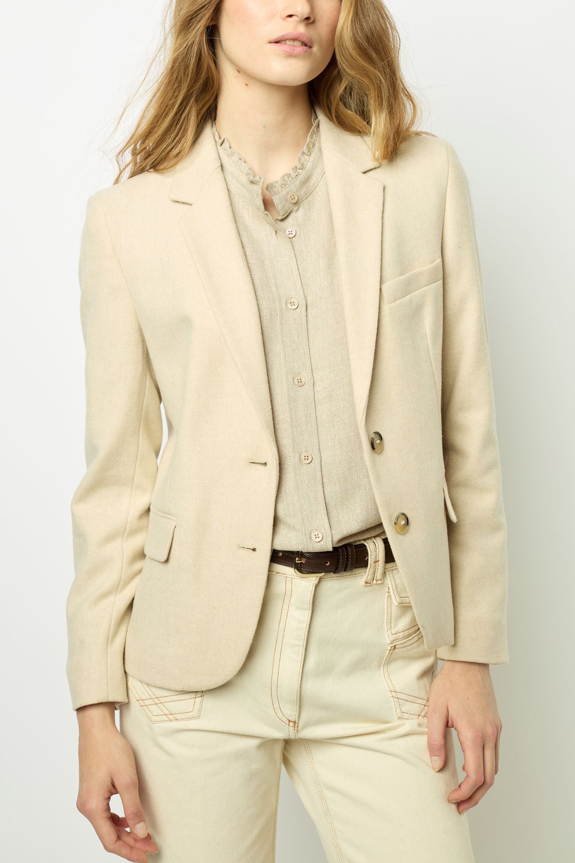 A woman wears a beige blazer over a light linen shirt, paired with cream-colored pants. She stands against a plain background, with her left hand resting at her side and her right hand in her pocket.