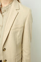 A person wearing a beige blazer over a light textured blouse with a ruffled collar, shown from the shoulders to the waist against a plain background.