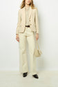 A person wearing a beige blazer, matching shirt, wide-leg cream pants, and black shoes holds a small beige textured handbag with a gold chain strap, standing against a plain white background.