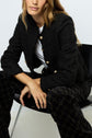 A woman sits on a black chair, wearing a textured black jacket with gold buttons, a white shirt, and black pants with a circular pattern. Her hair is wavy and brown.