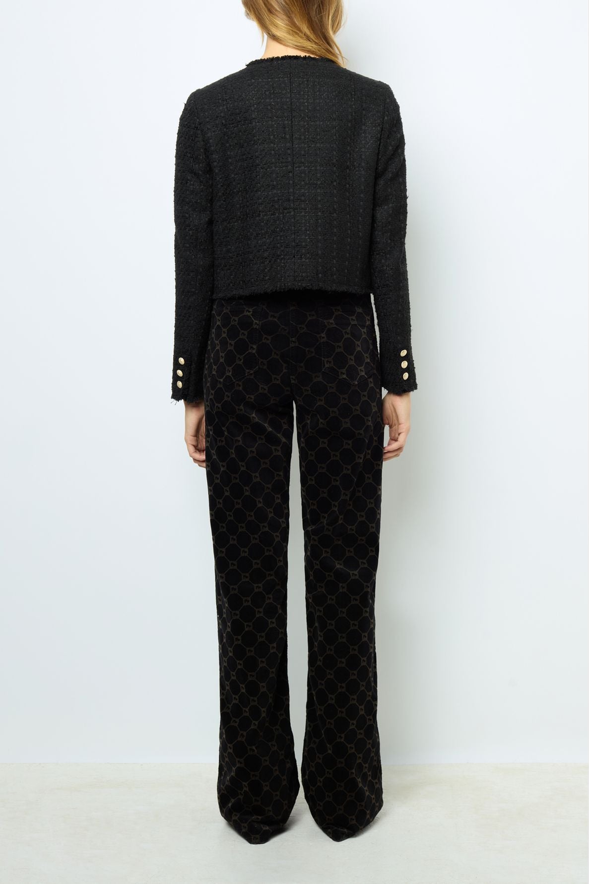 A person stands facing away, wearing a black textured jacket with gold buttons on the sleeves and black patterned pants. The background is plain white.