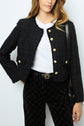 Woman wearing a textured black jacket with gold buttons over a white t-shirt and black patterned pants, carrying a black shoulder bag. Only her upper body is visible.
