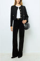 Woman wearing a black textured jacket with gold buttons, white top, black patterned pants, black shoes, and carrying a black shoulder bag, standing against a plain light background.