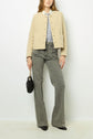 A woman stands against a white wall, wearing a beige textured jacket, light blue shirt, gray high-waisted flared jeans, black pointed shoes, and holding a small black handbag with a chain strap.