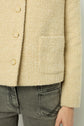 A close-up of a person wearing a textured beige jacket with gold buttons and a front pocket, paired with gray jeans that have visible stitching on the back pocket.