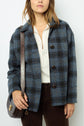 Woman wearing a veste esprit anglais—a blue veste à carreaux in laine vierge—over a white shirt, with brown corduroy pants and a brown shoulder bag, standing against a plain background.