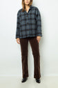 A person wearing a blue plaid button-up jacket in the style of a veste esprit anglais, brown corduroy pants, and black shoes stands against a plain white background.