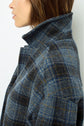 A person with shoulder-length brown hair wears a high-collared, blue and gray veste à carreaux in a classic esprit anglais style. The image shows a close-up side profile from the shoulders up against a white background.