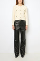 A woman stands against a white wall wearing a cream-colored buttoned jacket and black leather pants with black shoes. Her hair is down, and her arms are relaxed at her sides.