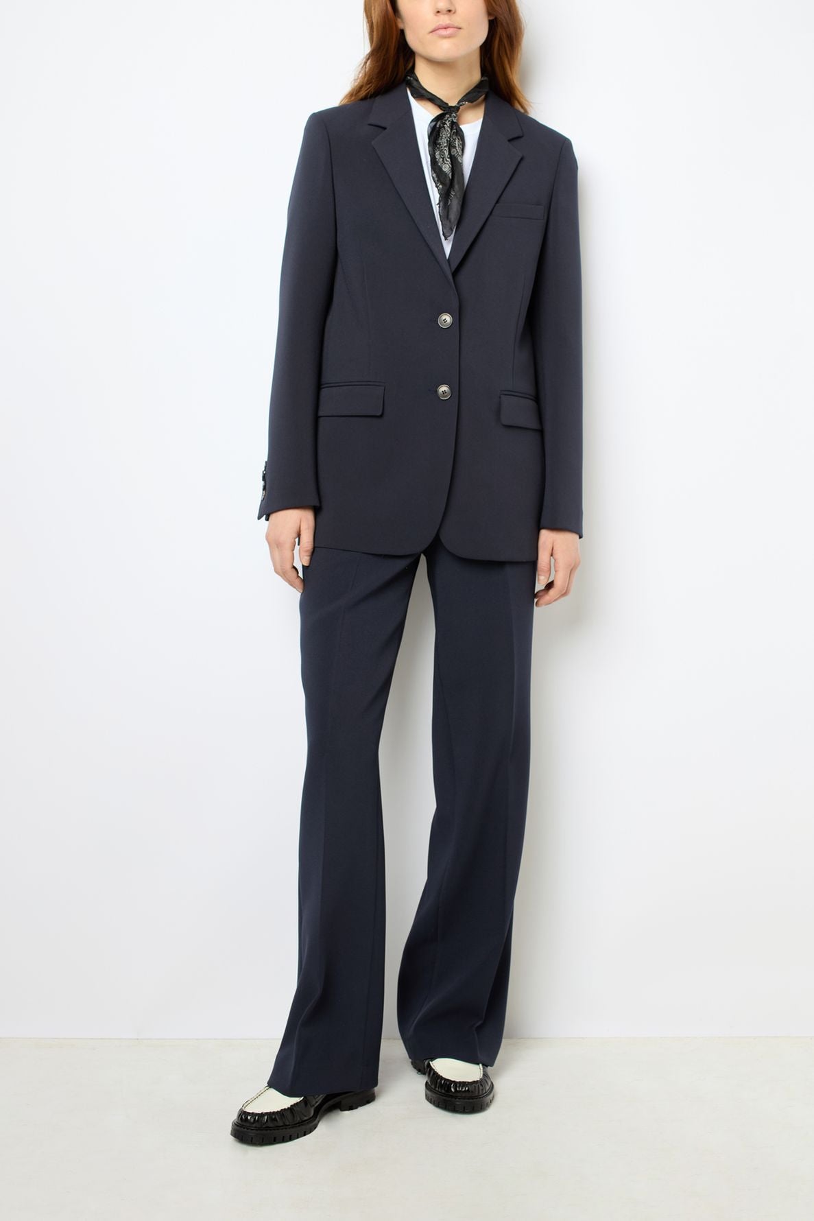 A person wearing a dark veste tailleur femme pantsuit with a white shirt, black neck scarf, and black-and-white shoes stands against a plain white background. The person’s head is partially out of frame.