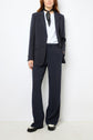 Woman wearing a veste tailleur femme, styled as a dark pantsuit with a white shirt, black scarf around her neck, and black shoes, standing against a plain white background.