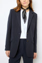 A person wearing a navy blue veste tailleur femme and trousers, a crisp white shirt, and a black patterned scarf around their neck, standing against a plain white background.