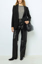 A person wearing a veste noire, black and white striped shirt, black leather pants, black pointed shoes, and carrying a black handbag stands against a plain light background.