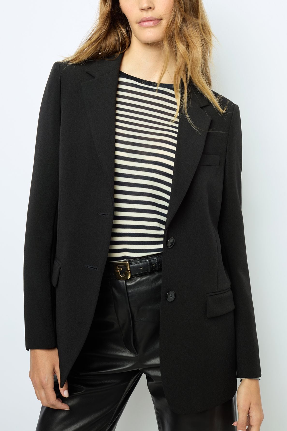 Woman wearing a veste tailleur femme noire over a black and white striped shirt with black leather pants. Her hair is loose, and she is standing against a plain white background. Her face is partially out of the frame.