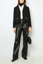 Woman in black microfibre noir leather pants, white shirt, black veste courte cropped blazer with chaînes dorées gold chains, black turtleneck, black shoes, and holding a small black bag with a gold chain handle, standing against a white background.