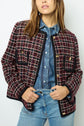 A woman wearing a red, black, and white plaid veste tweed over a blue denim shirt and light blue jeans, standing against a plain light background.