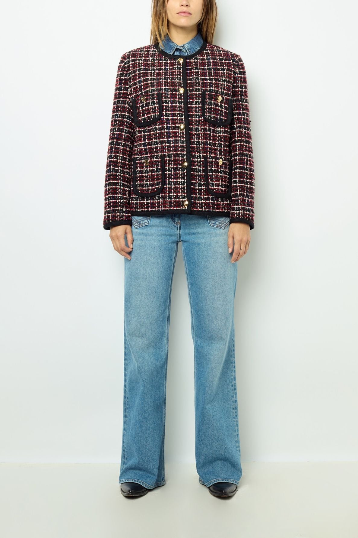 A person stands against a white wall wearing a veste rouge noir in a red, black, and white plaid pattern with front pockets, a blue shirt underneath, light blue wide-leg jeans, and black shoes.