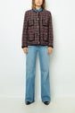 A person stands against a white wall wearing a veste rouge noir in a red, black, and white plaid pattern with front pockets, a blue shirt underneath, light blue wide-leg jeans, and black shoes.