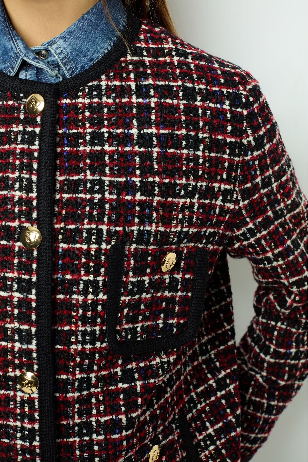 Close-up of a person wearing a red, black, and white plaid textured veste tweed sans col with gold buttons over a blue denim shirt. The jacket features a decorative front pocket with a matching gold button.