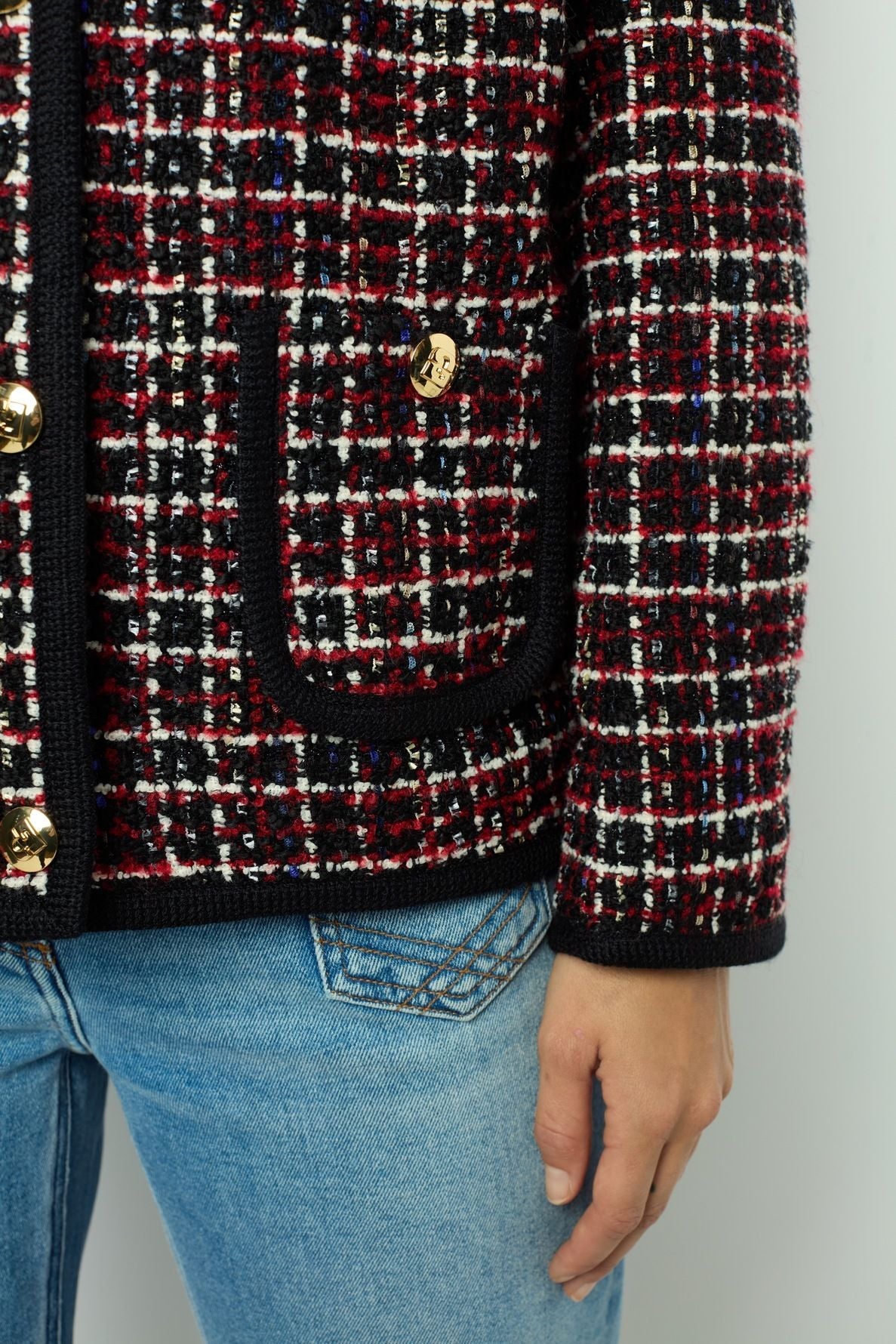 A person wearing light blue jeans and a red, black, and white checkered veste tweed with gold buttons, showing only the lower torso and one hand.