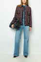 A woman wearing a red and black checked veste tweed jacket, blue denim shirt, light blue wide-leg jeans, and black shoes, holding a large black handbag, stands against a plain white background.