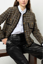 Woman wearing a white button-up shirt, a brown and black veste en tweed with gold buttons, and black leather pants sits on a wooden bench against a plain background.