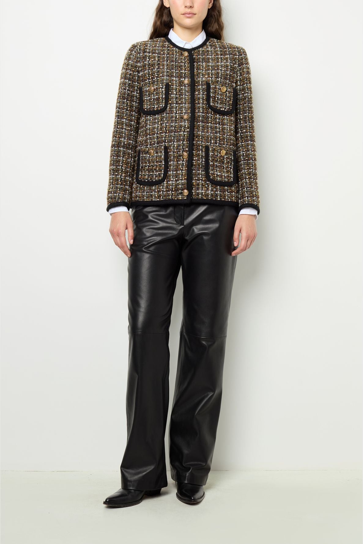 A person stands against a white wall wearing a brown plaid veste sans col with front pockets and black trim, paired with black leather pants and black shoes. Their hands are relaxed at their sides.