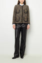 A person stands against a white wall wearing a brown plaid veste sans col with front pockets and black trim, paired with black leather pants and black shoes. Their hands are relaxed at their sides.