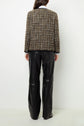 A person stands facing away, wearing a brown and white veste en tweed textured jacket and black leather pants. Their hair is long and dark, and they are posed against a plain white background.