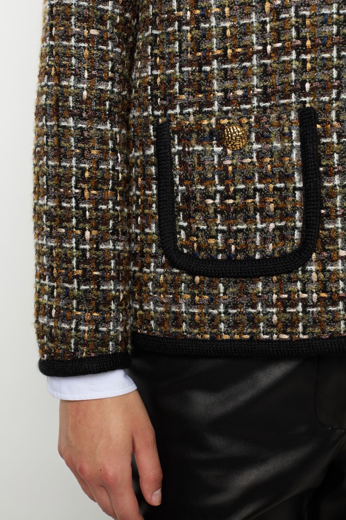 A close-up of a person wearing a veste en tweed with a black-trimmed pocket and gold button, over a white cuffed shirt and black pants.