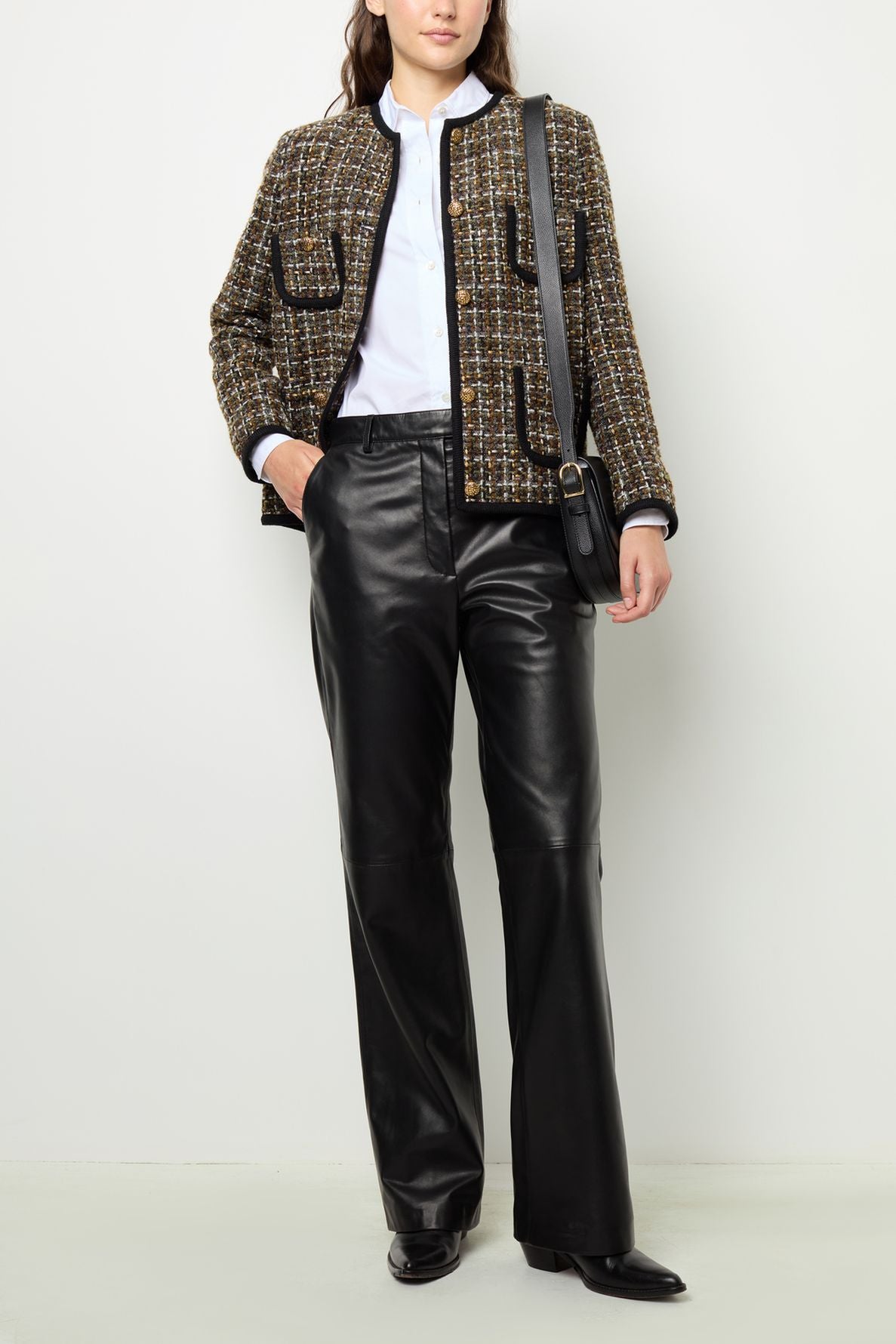Woman wearing a veste en tweed, white shirt, black leather pants, black shoes, and a black shoulder bag, standing against a plain white background.