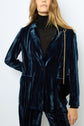 Woman wearing a dark blue velvet suit with a black turtleneck and a small black purse with a gold chain shoulder strap, standing against a plain light background.