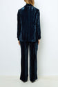 A person standing with their back to the camera, wearing a dark blue velvet suit with long sleeves and wide-leg pants, against a plain white background.
