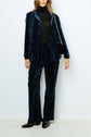 Person wearing a dark blue velvet suit with a black turtleneck, black heeled boots, and a small purse with a chain strap, standing against a plain white background.