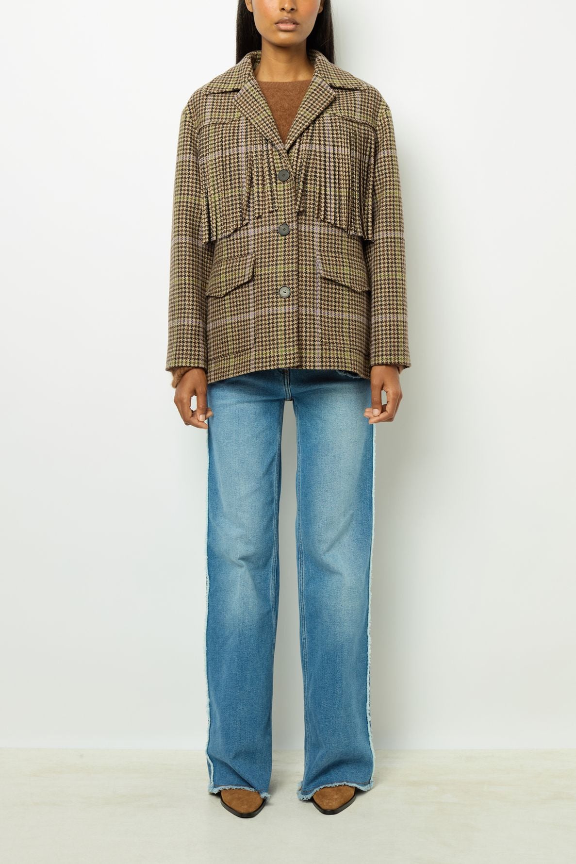 A person stands against a white background wearing a brown plaid blazer en laine, a brown top, blue flared jeans, and brown pointed shoes. The photo is cropped just above the eyes.