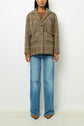 A person stands against a white background wearing a brown plaid blazer en laine, a brown top, blue flared jeans, and brown pointed shoes. The photo is cropped just above the eyes.