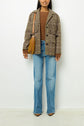 Woman wearing a brown laine sweater, plaid jacket with an imprimé Pied de Poule pattern, blue jeans, and pointed-toe shoes, carrying a mustard-yellow handbag, standing against a plain white background.