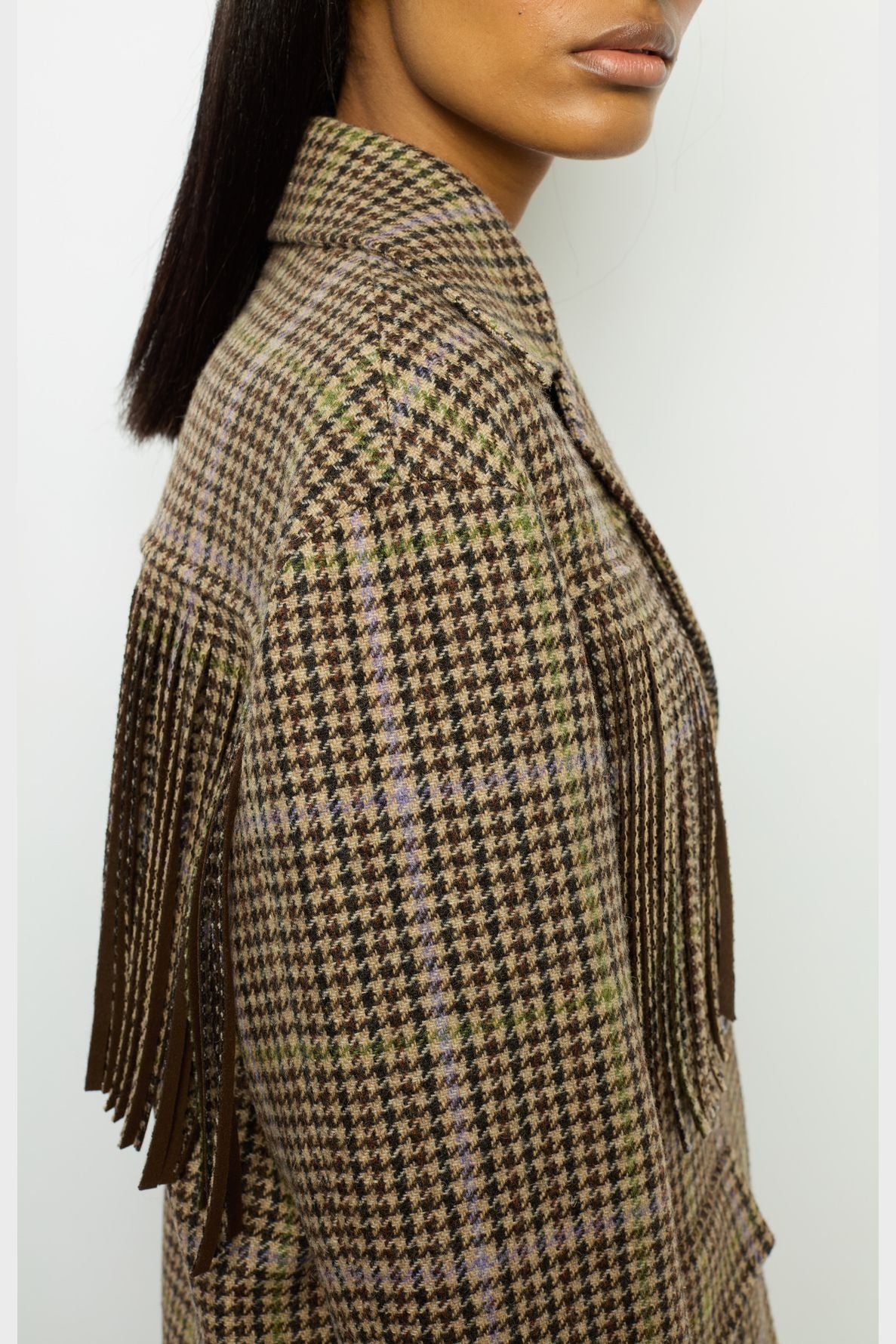 A woman wearing a brown and green plaid jacket crafted from laine, with long fringe detailing on the shoulder in the style of a veste à franges, shown from the side against a plain background.