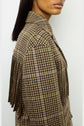 A woman wearing a brown and green plaid jacket crafted from laine, with long fringe detailing on the shoulder in the style of a veste à franges, shown from the side against a plain background.