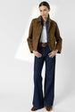 Woman standing against a white background, wearing a brown cuir velours veste courte femme, a white shirt, blue denim jeans, a belt, brown shoes, and layered necklaces. She has her hands in her pockets and her hair pulled back.