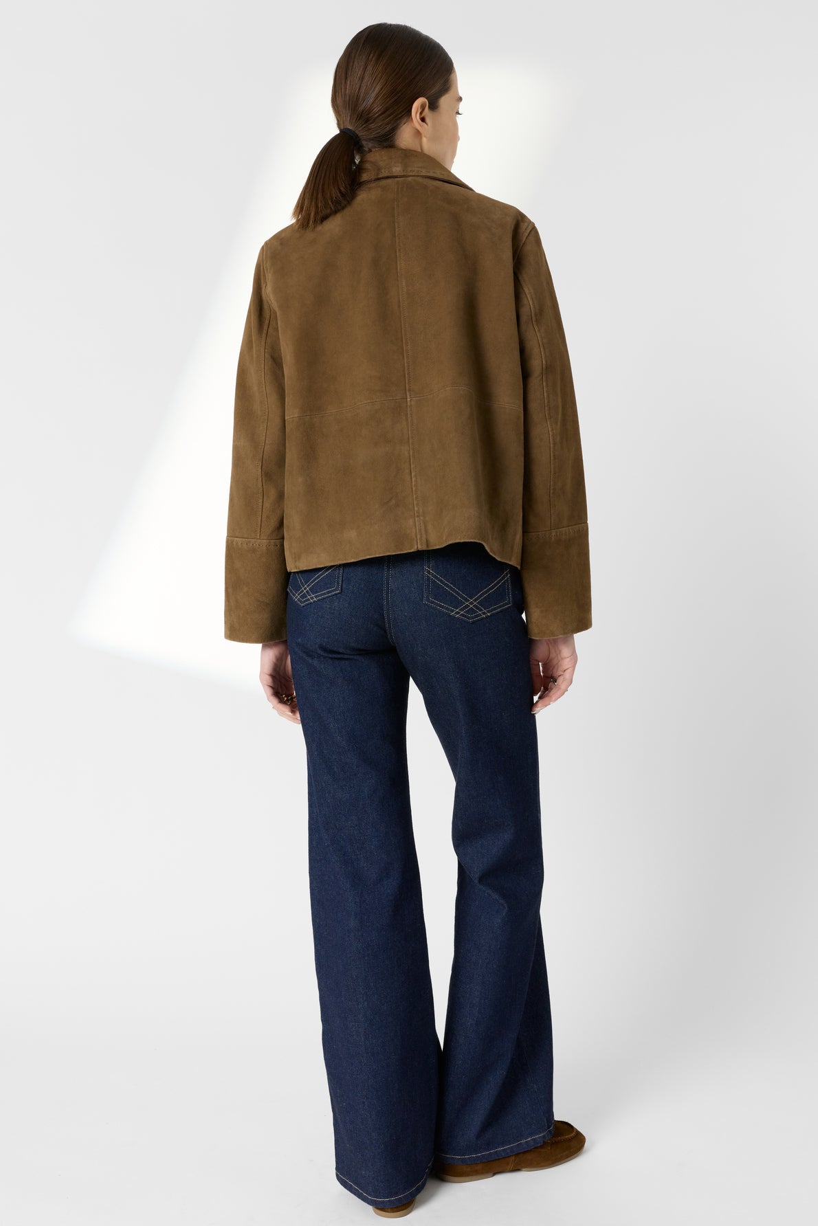 A person stands facing away, wearing a brown veste en cuir velours, dark blue flared jeans, and brown shoes. The background is plain and light-colored.