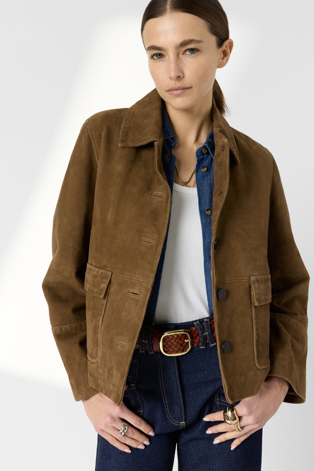 A woman with straight brown hair wears a brown suede cuir velours veste courte femme over a blue shirt and white top, paired with dark jeans and a braided leather belt. She stands with one hand in her pocket against a light background.