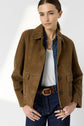 A woman with straight brown hair wears a brown suede cuir velours veste courte femme over a blue shirt and white top, paired with dark jeans and a braided leather belt. She stands with one hand in her pocket against a light background.