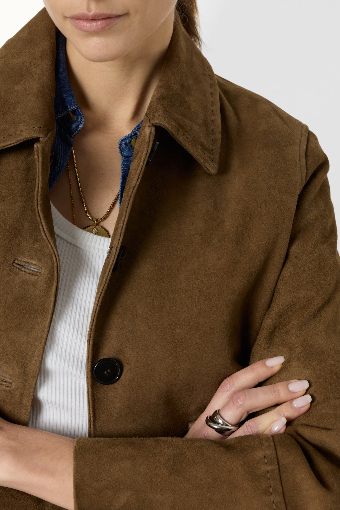 A woman wears a veste courte femme in brown cuir velours over a white top and blue shirt, accessorized with a gold necklace and a chunky ring, arms crossed.