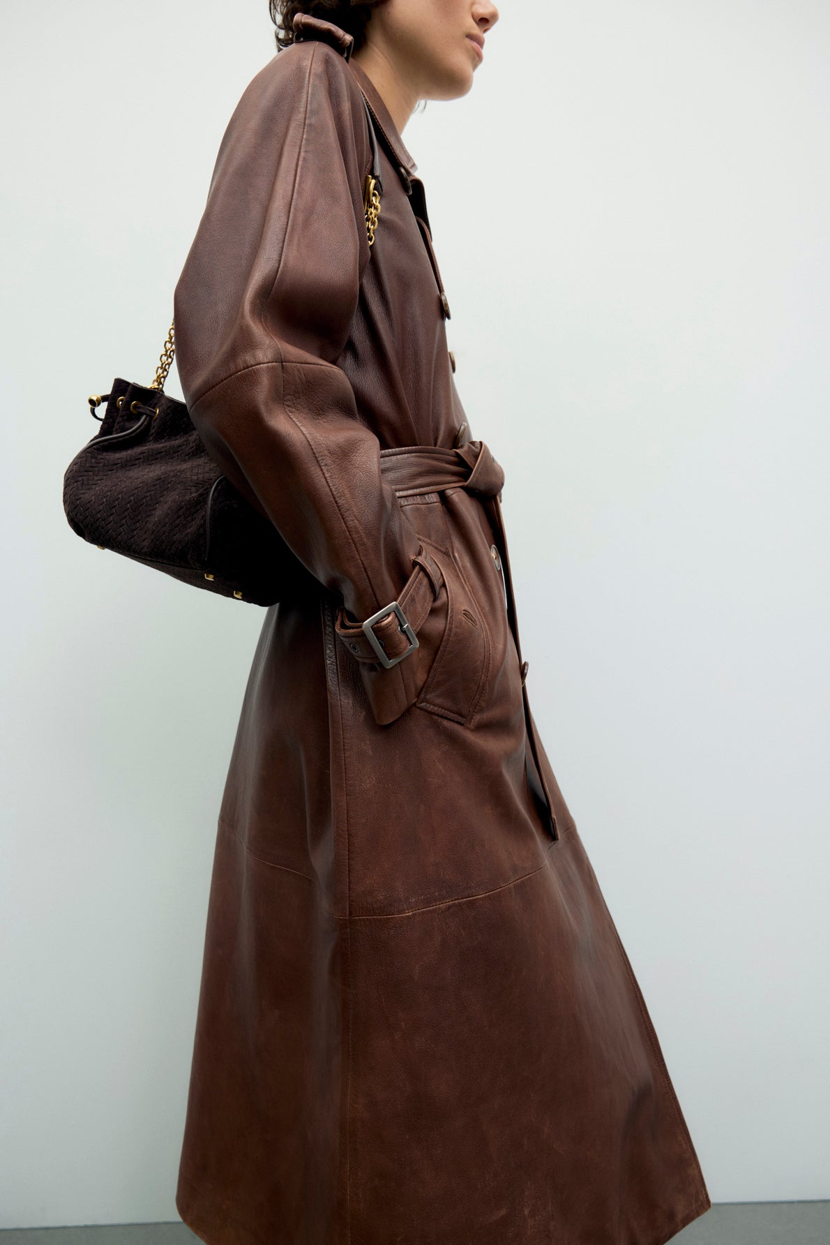 Person wearing a long brown trench-coat cuir with a belt and side pockets, carrying a black shoulder bag with a gold chain strap, standing against a plain light background. This manteau femme effortlessly combines timeless elegance and modern style.