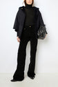 A person wearing a black turtleneck, black flared pants with a subtle pattern, black shoes, a luxurious cape peau lainée coat, and holding a black handbag, standing against a white background.