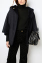 Woman in a black turtleneck, black textured pants, and a black hooded manteau en mouton holds a black leather bucket bag. She stands against a plain white background.