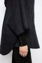 A person wearing a dark, oversized manteau en mouton with a fuzzy inner lining, dark patterned pants, and a black sweater with button details on the sleeve stands against a plain light background.