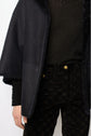 Person wearing a black cape peau lainée with a furry collar, black top, and black patterned pants with gold buttons, showing only the torso and part of the left arm.